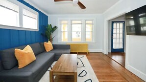 Ein Wohnzimmer mit einem dunklen Sofa, einem Holztisch, einem Deckenventilator und großen Fenstern mit Jalousien.