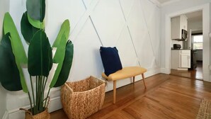 Ein Zimmer mit Holzboden, einem gewebten Korb, einem Stuhl und großen, grünen Blättern an einer weißen Wand.