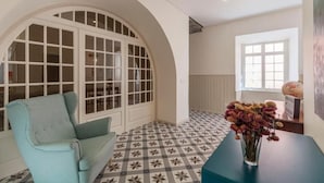 A room with a patterned floor, a light blue armchair, a vase with flowers, and a large arched window. - A Bica Hostel (Ponta Delgada)