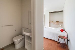 A small, modern bathroom with a toilet, sink, and mirror. There is a bed with a white sheet and a red lamp on a wooden chair. - A Bica Hostel (Ponta Delgada)