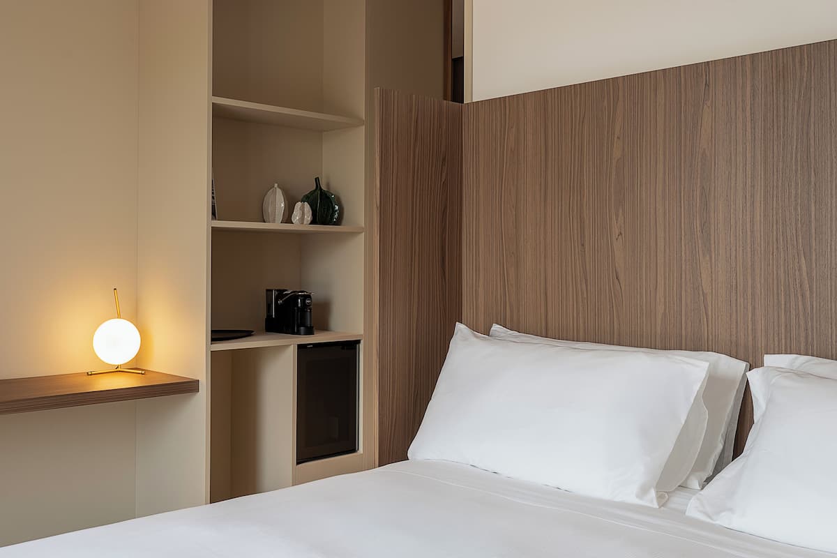 Habitación de hotel con cama, mesita de noche con lámpara, repisa con adornos y cabecero de madera.