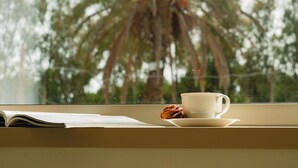 Una ventana con una taza y un libro sobre una mesa, una palmera y un cielo despejado.