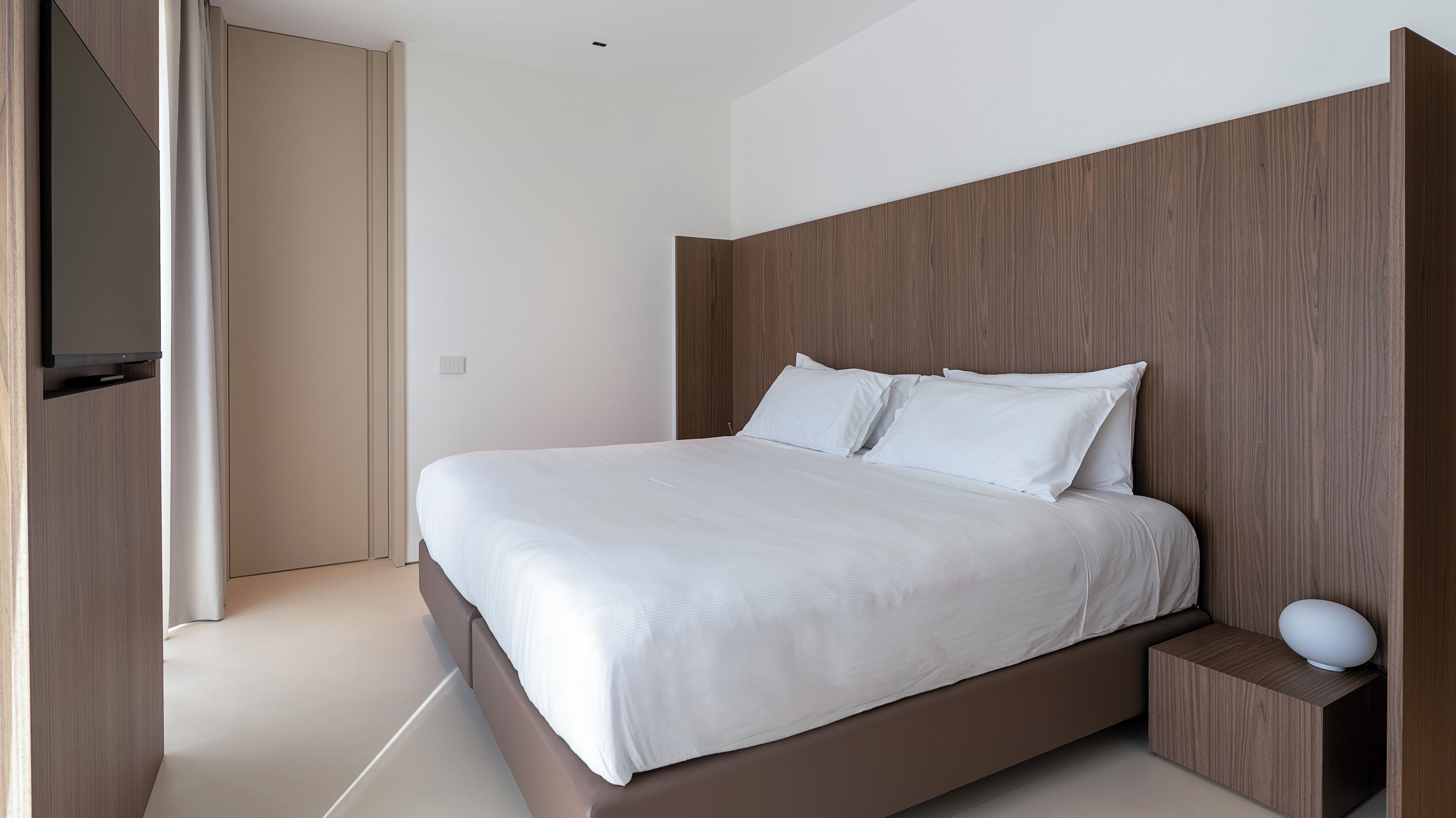 Un dormitorio moderno con una cama grande, cabecera de madera, una mesita de noche y un televisor montado en la pared.
