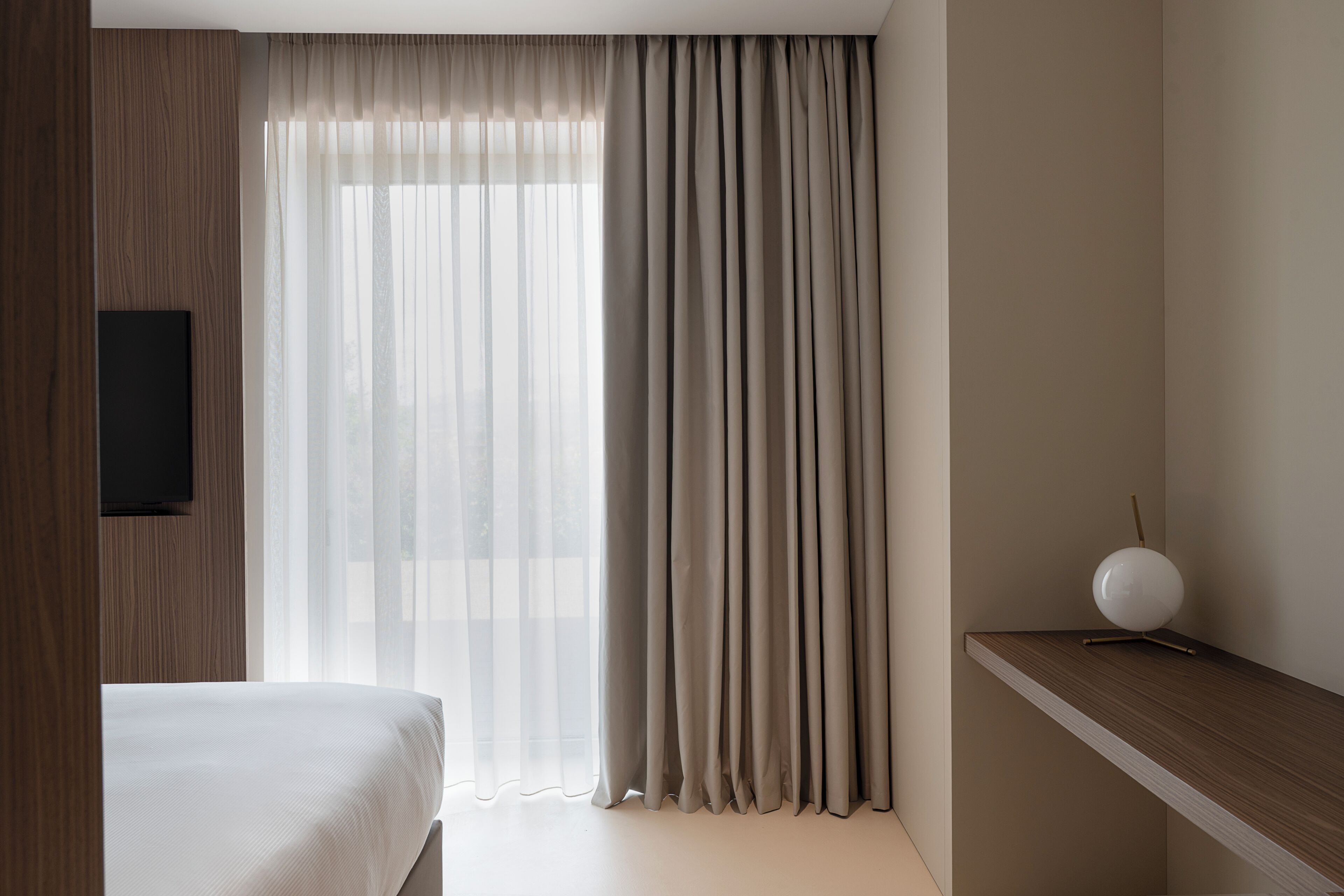 Ein Hotelzimmer mit einem Bett, einem an der Wand befestigten Fernseher, einem Fenster mit leichtem Vorhang und einem Nachttisch mit Lampe.