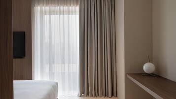 Ein Hotelzimmer mit einem Bett, einem an der Wand befestigten Fernseher, einem Fenster mit leichtem Vorhang und einem Nachttisch mit Lampe.