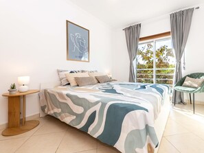 Ein Schlafzimmer mit Bett, Nachttischen, einem Sessel, einem Fenster mit Vorhängen und einem gerahmten Kunstwerk an der Wand.