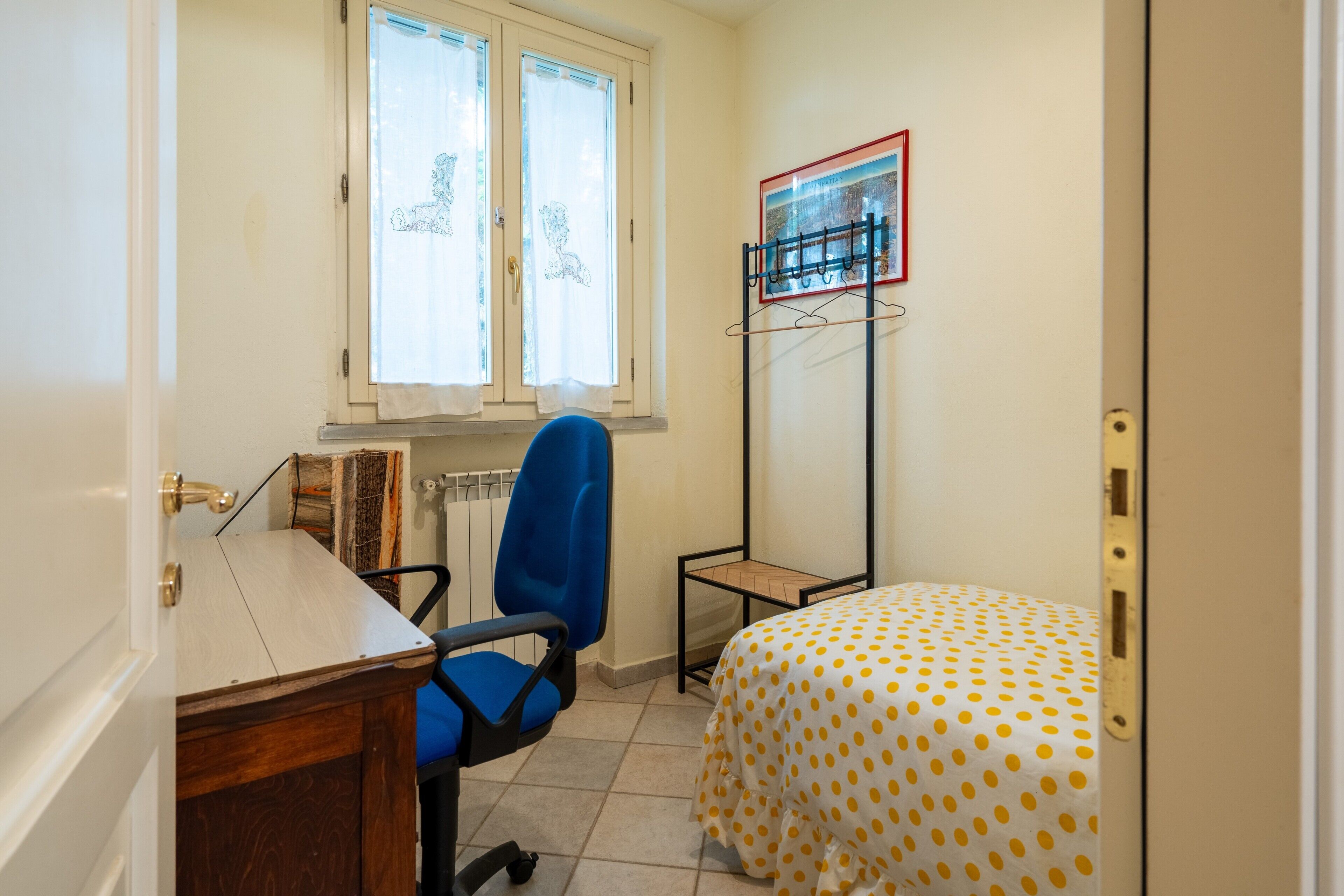 A room with a desk, a blue chair, a bed with a polka dot bedspread, a radiator, and a window with curtains.