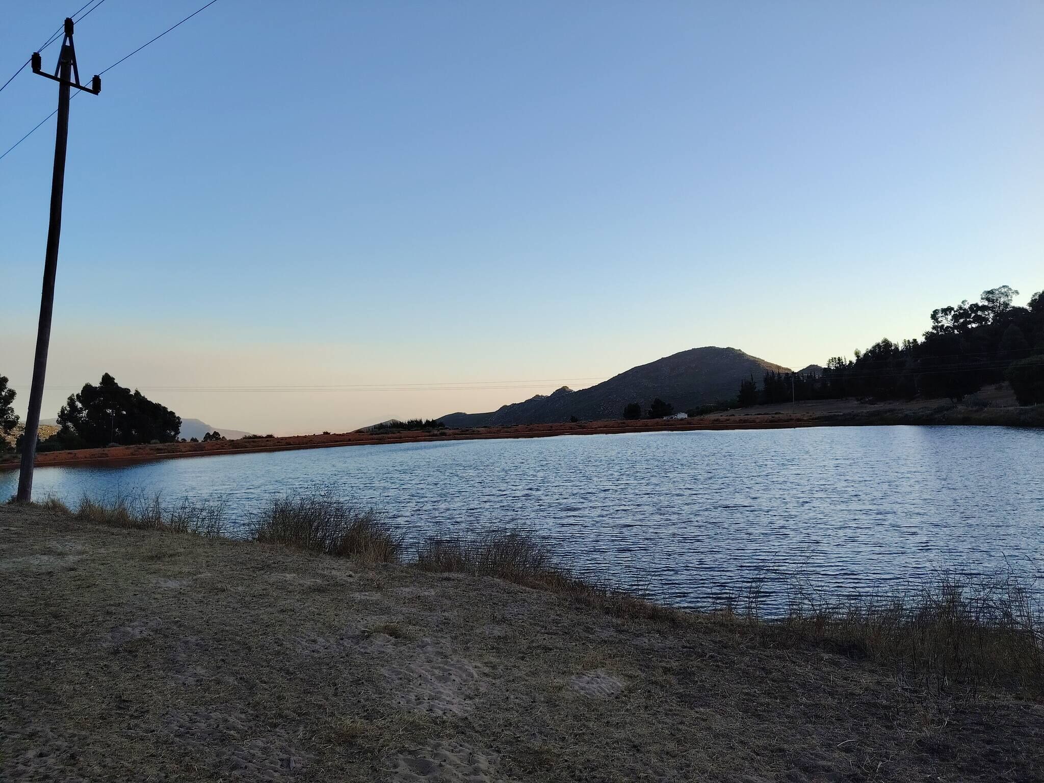 Un lago tranquilo con una montaña al fondo, rodeado de vegetación escasa y un poste de electricidad.