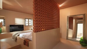A bedroom with a TV, a bed, a window, and a mirror. - Terra Verde Oia Suites - Aurea Suite Sea View with Outdoor Jetted Tub (Oia)