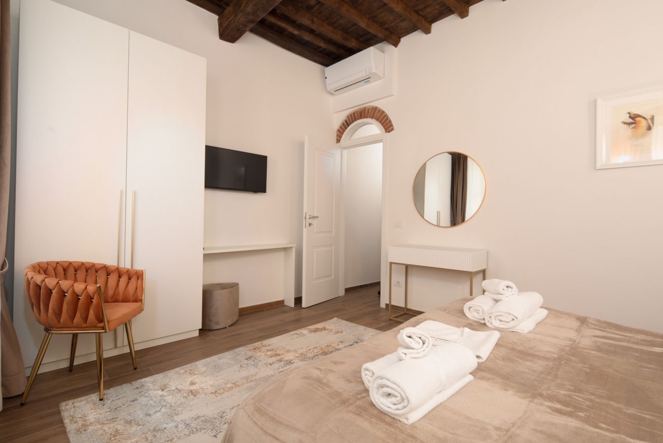 A bedroom with a bed, a chair, a vanity, a mirror, and a television.