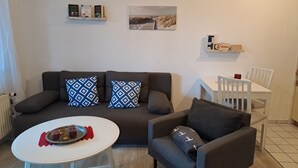 A living room with a sofa, armchair, and a small table with drinks. There is a painting on the wall and a shelf with books.