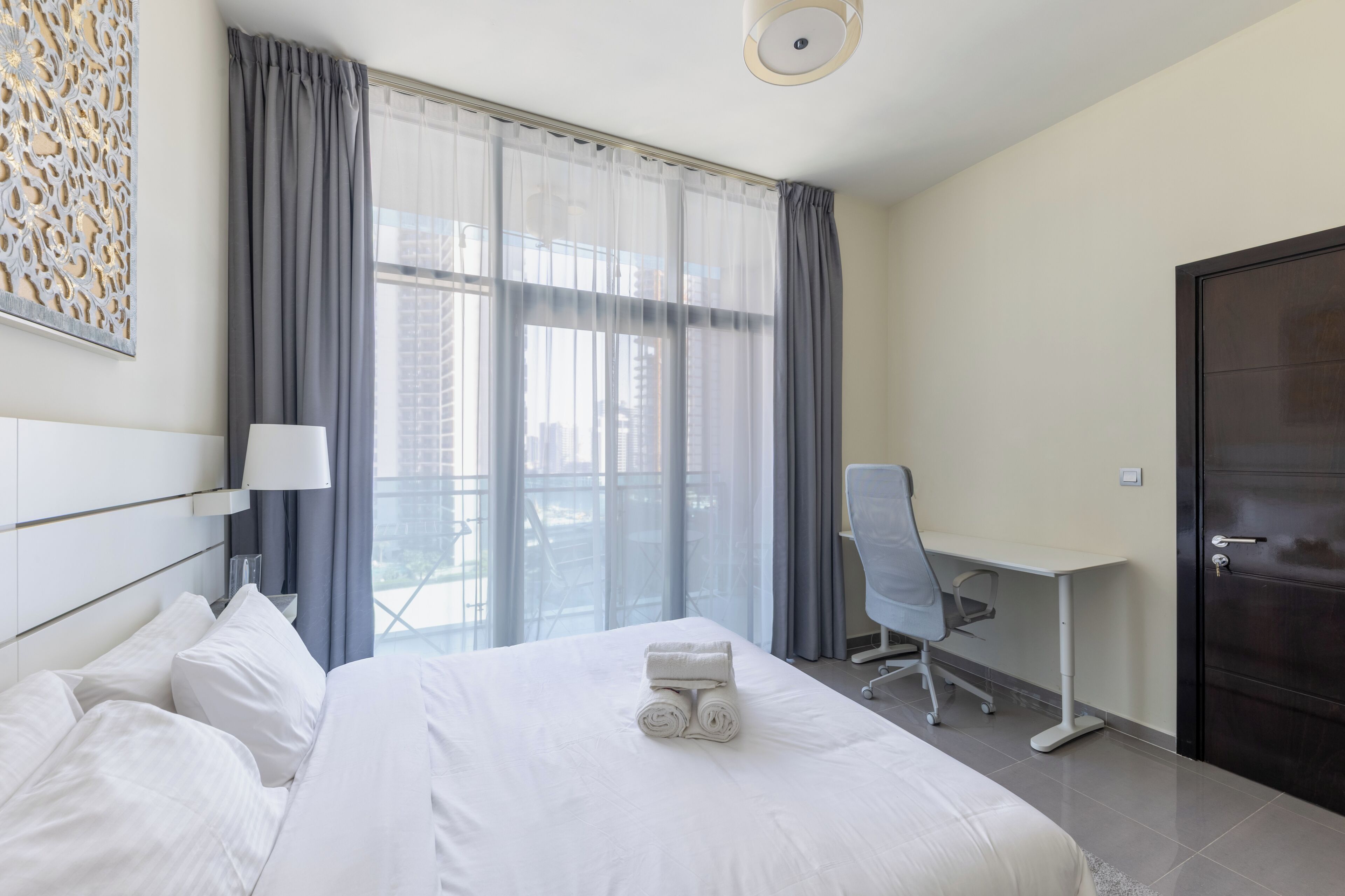A hotel room with a bed, a desk, a chair, a window with curtains, and a door.