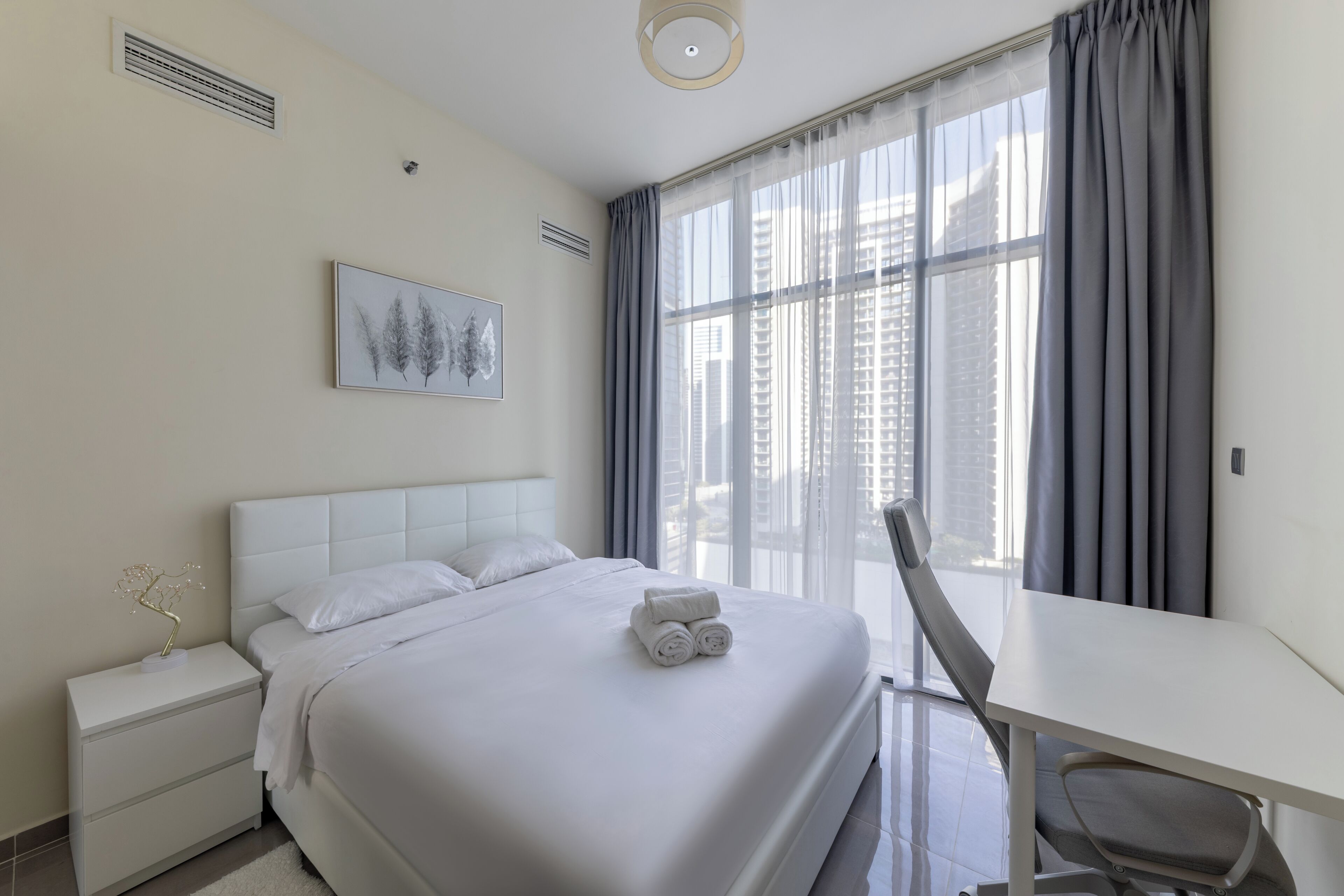 A hotel room with a bed, a desk, a chair, a nightstand, a picture on the wall, and a window with curtains.