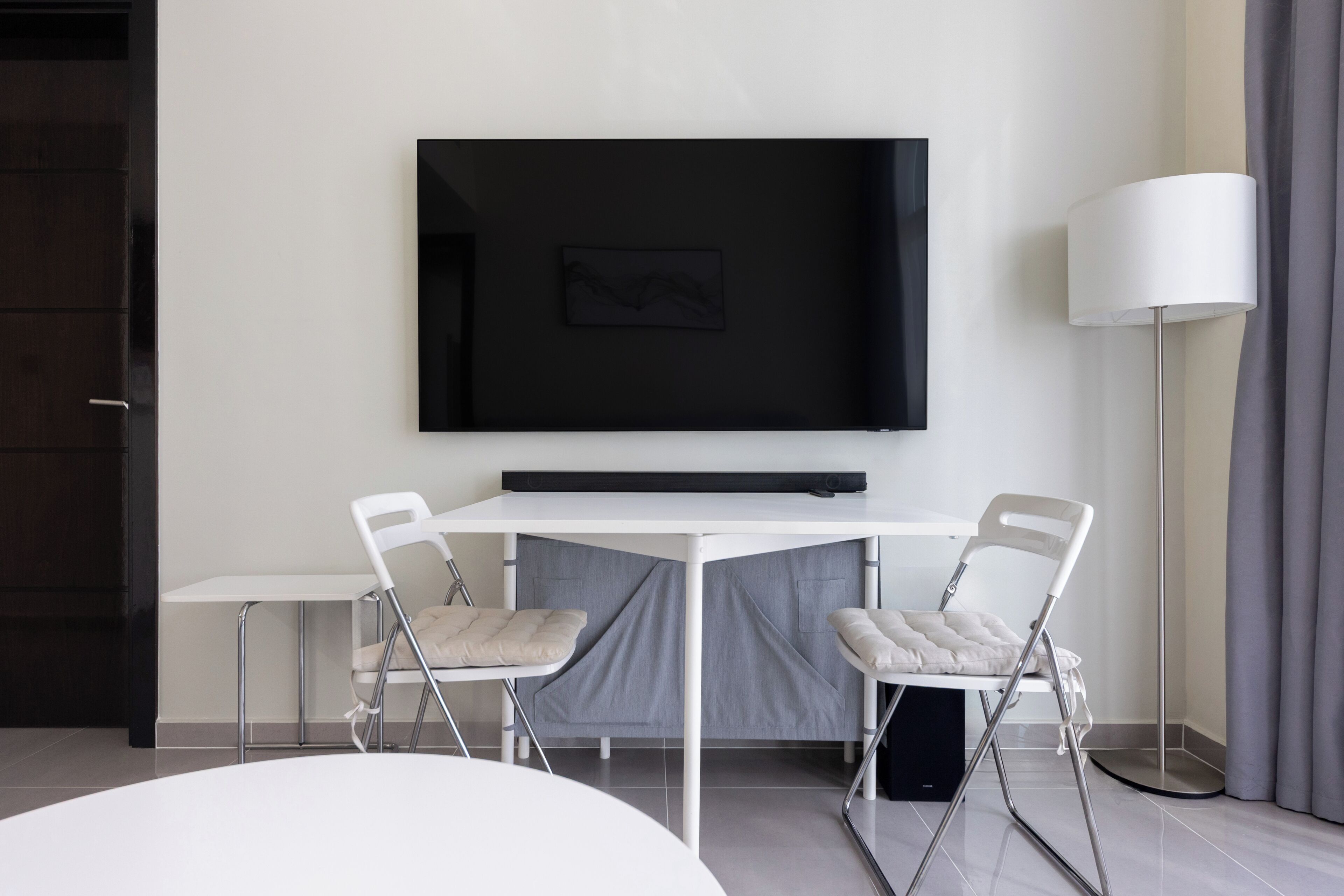 Ein modernes Hotelzimmer mit einem an der Wand befestigten Flachbildfernseher, einem weißen Esstisch mit zwei Stühlen, einer Stehlampe und einem großen Fenster mit Vorhängen.