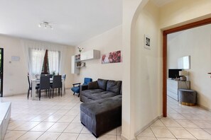 Un soggiorno moderno con un divano grigio, un'area pranzo con tavolo e sedie, e una cucina con TV e lampada.