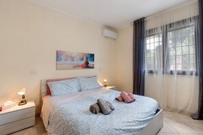 A hotel room with a bed, a nightstand, a wall-mounted air conditioner, and a window with curtains.