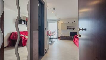 An open door leading to a modern living room with a dining area, a TV, and a red-covered sofa.