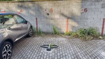 Une voiture est garée à côté d’un mur divisé en sections numérotées, un tuyau d’arrosage est posé sur le sol et le sol est pavé de briques.