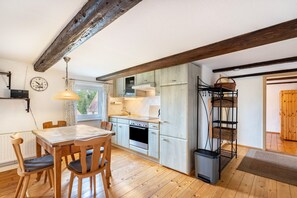A kitchen with wooden cabinets, a dining table with chairs, a hanging light fixture, and a window with curtains. - Holiday Apartment for 3 Guests (45 m²) in Bertsdorf-Hörnitz (Bertsdorf-Hörnitz)