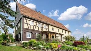 A traditional half-timbered house with a stone foundation, a tiled roof, and a well-maintained garden with various plants and a stone wall. - Koitsche Apartment for 3 Guests (35 m²) in Bertsdorf-Hörnitz (Bertsdorf-Hörnitz)