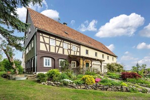 Une maison traditionnelle à colombages, avec des fondations en pierre, un toit en tuiles et un jardin bien entretenu, agrémenté de diverses plantes et d’un mur en pierre.