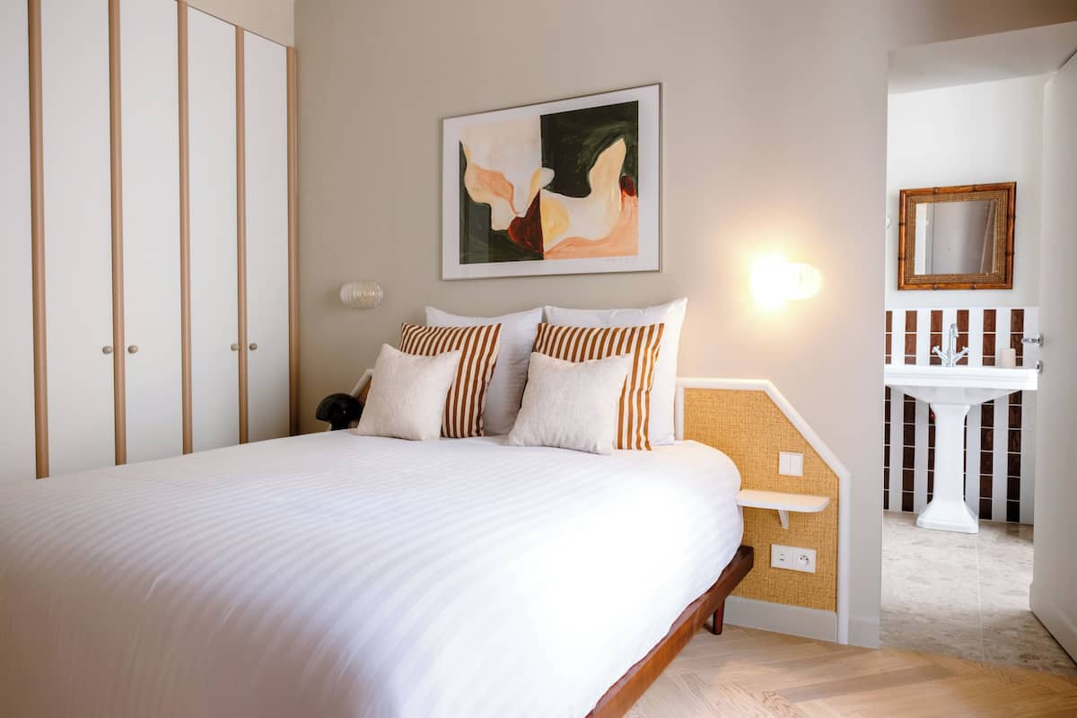 A bedroom with a bed, a painting, a bedside lamp, a mirror, and a bathroom with a sink and pedestal.