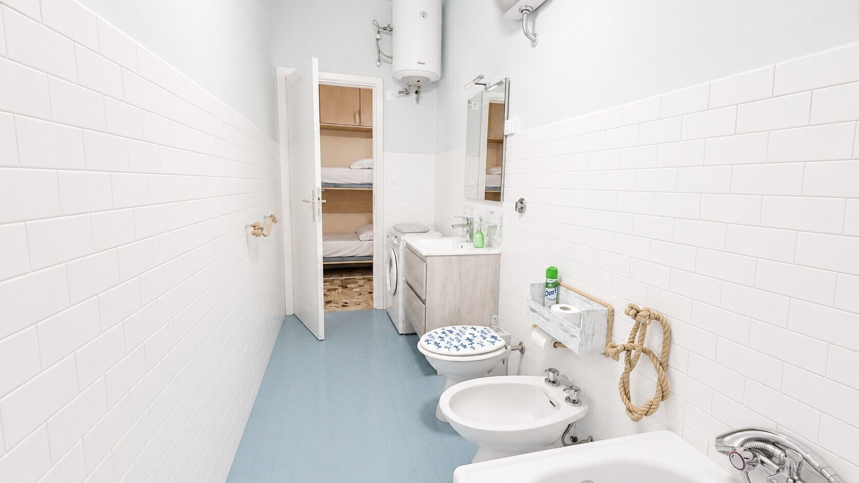 Ett litet badrum med tvättmaskin, toalett, handfat och en liten säng i bakgrunden.