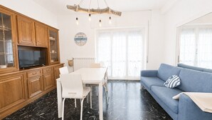 Un salon avec un canapé bleu, une table à manger avec des chaises, des meubles en bois, un téléviseur et une grande fenêtre avec des rideaux.