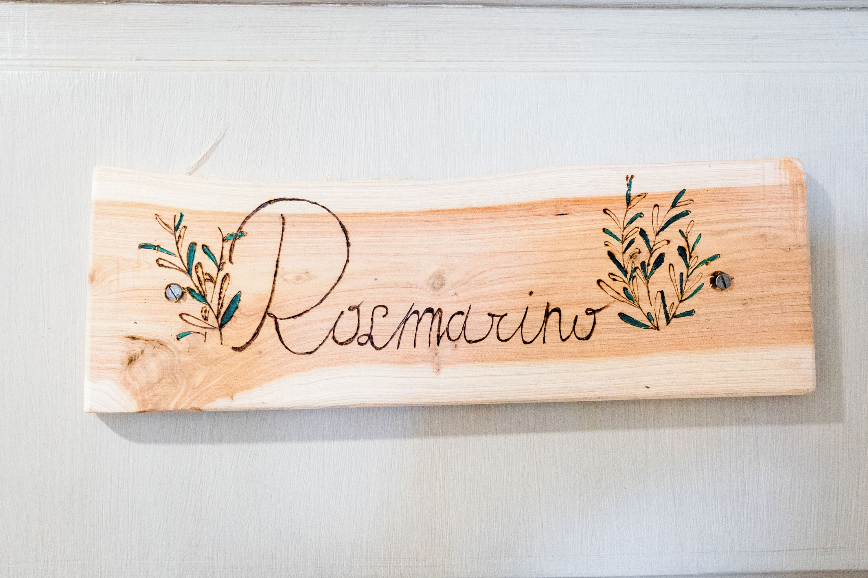 Ein hölzernes Schild mit dem Schriftzug 'Rosmarino' und dekorativem Blattwerk.