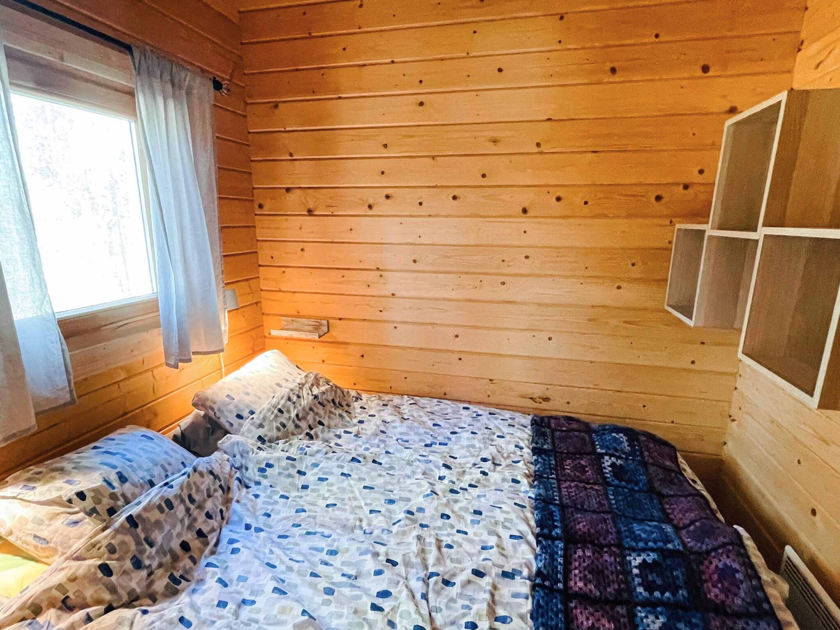 A wooden cabin room with a bed, a window with curtains, and a wall-mounted shelf.
