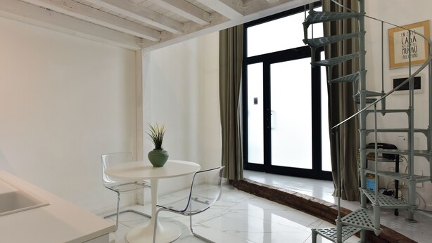 A modern room with a white table, clear chairs, a potted plant, a metal spiral staircase, and a large window with curtains. - 4BNB - Loft Colosseum 3 (Rome)