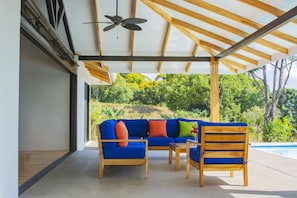 A covered outdoor seating area with a sofa and chairs, a ceiling fan, and a view of a swimming pool and greenery.