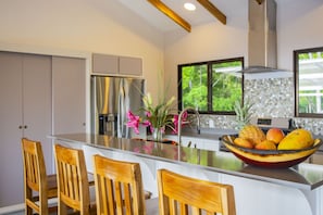 A modern kitchen with a central island, wooden chairs, a stainless steel refrigerator, and a fruit bowl.
