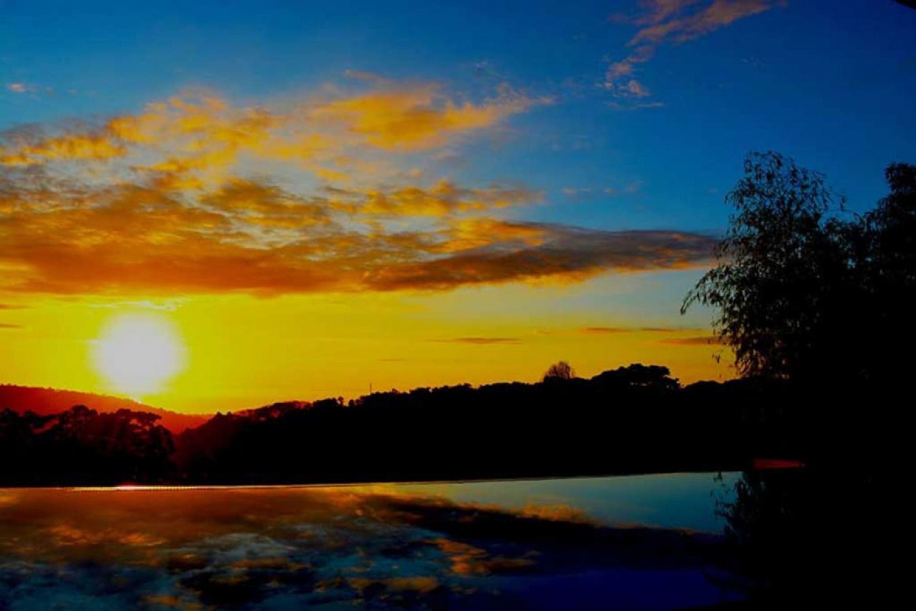 Ein Sonnenuntergang über einem Gewässer, im Hintergrund sind Baumreihen und ein Berg schattenhaft zu sehen.