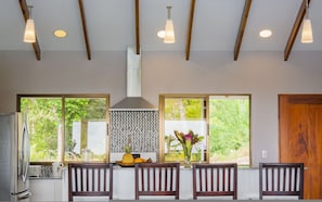 A modern kitchen with a large window, wooden ceiling beams, and a central island with chairs.