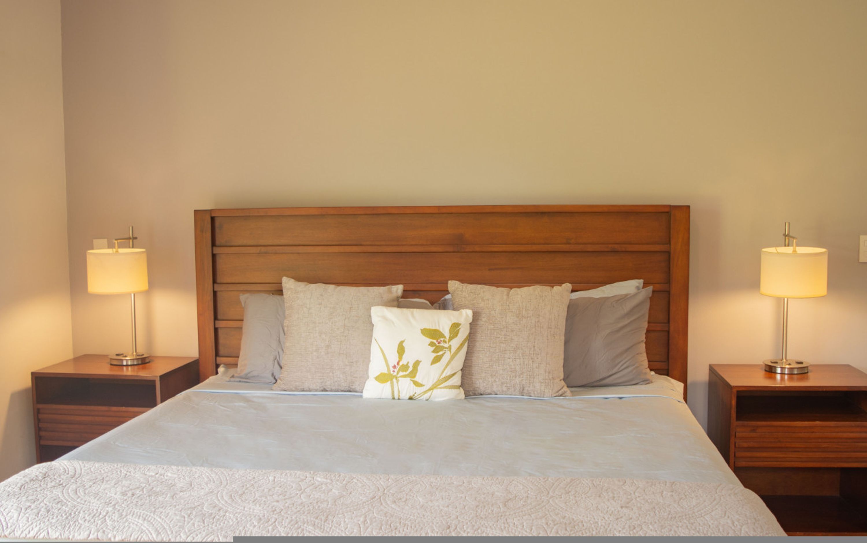 Un lit bien fait, avec une tête de lit en bois, deux tables de chevet avec des lampes et un coussin à motifs floraux.