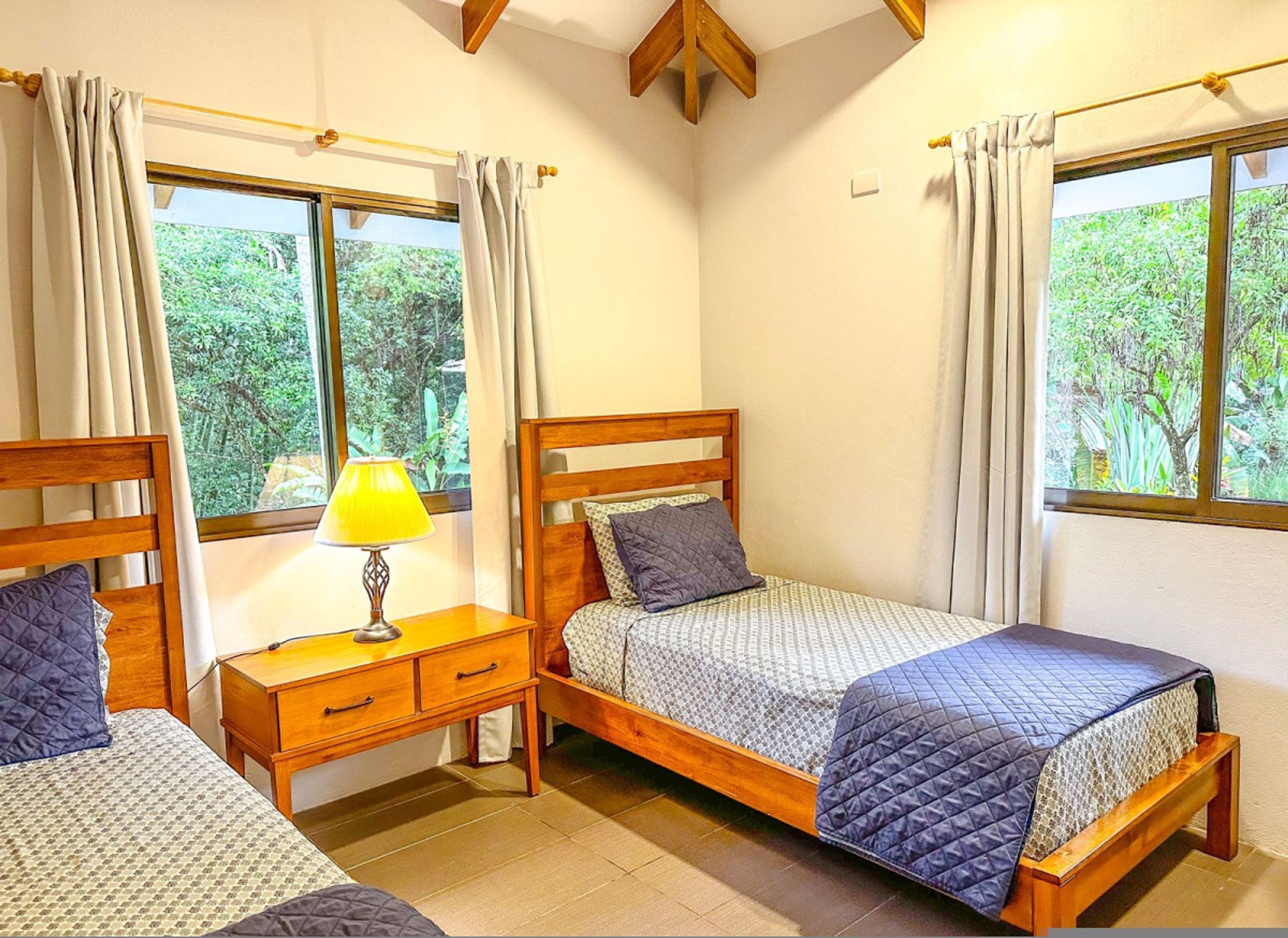A room with two single beds, a bedside table with a lamp, and a window with curtains.