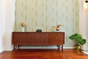 Ein Holzkabinent mit Schubladen, eine Schreibmaschine und ein Blumentopf vor einer Wand mit geometrischem Muster.