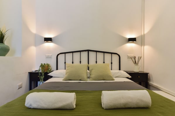 A neatly made bed with two pillows, a green bedspread, and two white towels. There are two bedside tables with lamps, a potted plant, and a vase with reeds. - 4BNB - Loft Colosseum 1 (Rome)