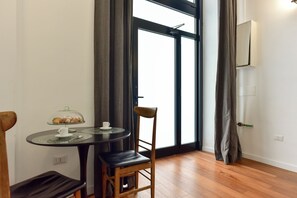 A room with a round table, a chair, a glass display case with pastries, and a large window with curtains. - 4BNB - Loft Colosseum 1 (Rome)