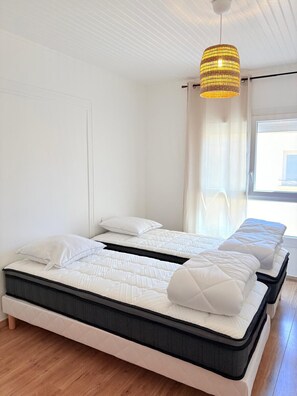 Ein modernes Schlafzimmer mit einem großen Bett, einem kleineren Bett, einer Hängeleuchte und einem Fenster mit Vorhängen.