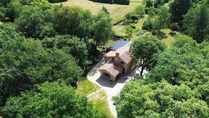 An aerial view of a house surrounded by dense greenery, with a paved driveway leading to the entrance.