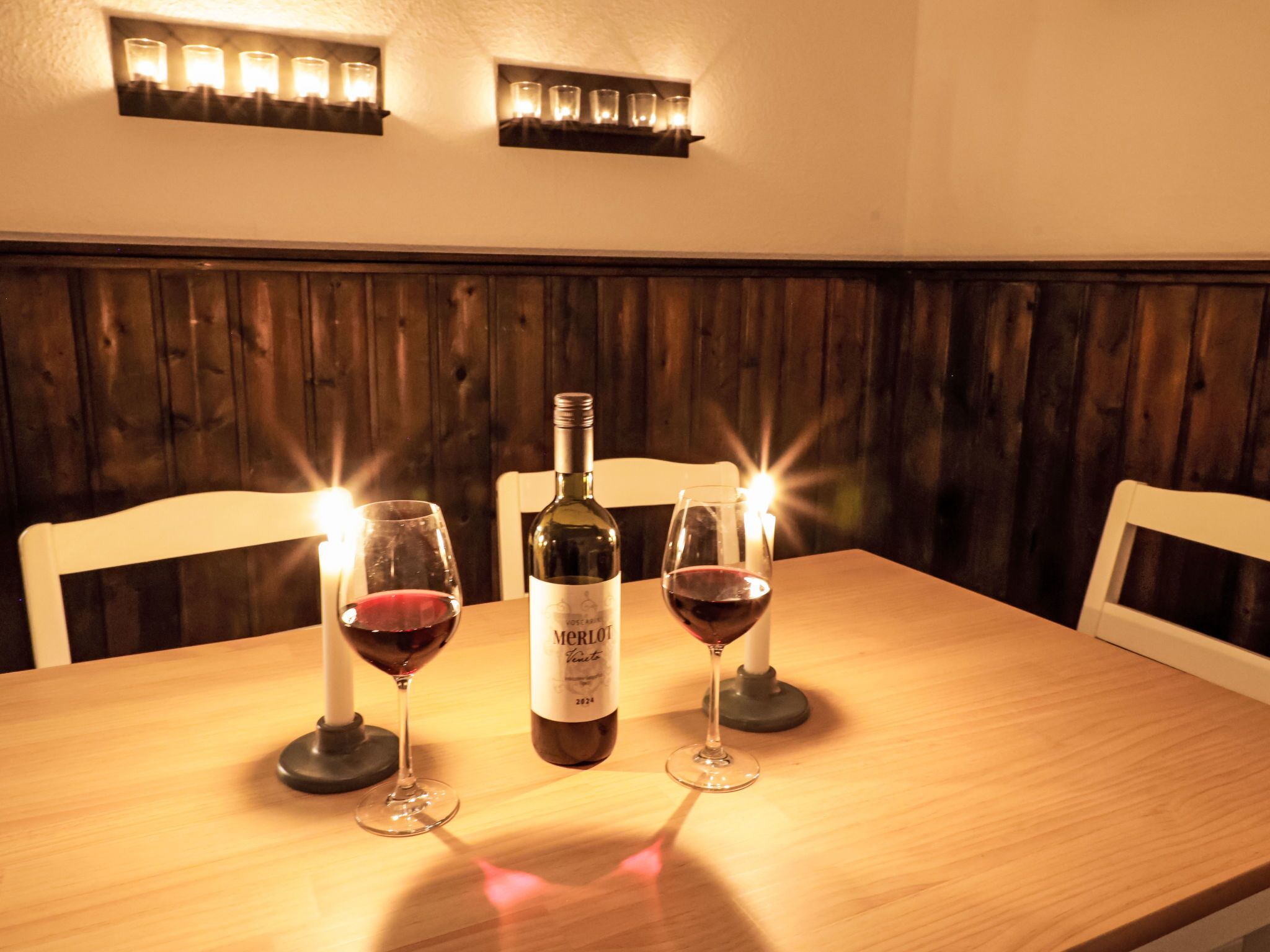 A wooden table with two wine glasses and a bottle of Merlot wine.