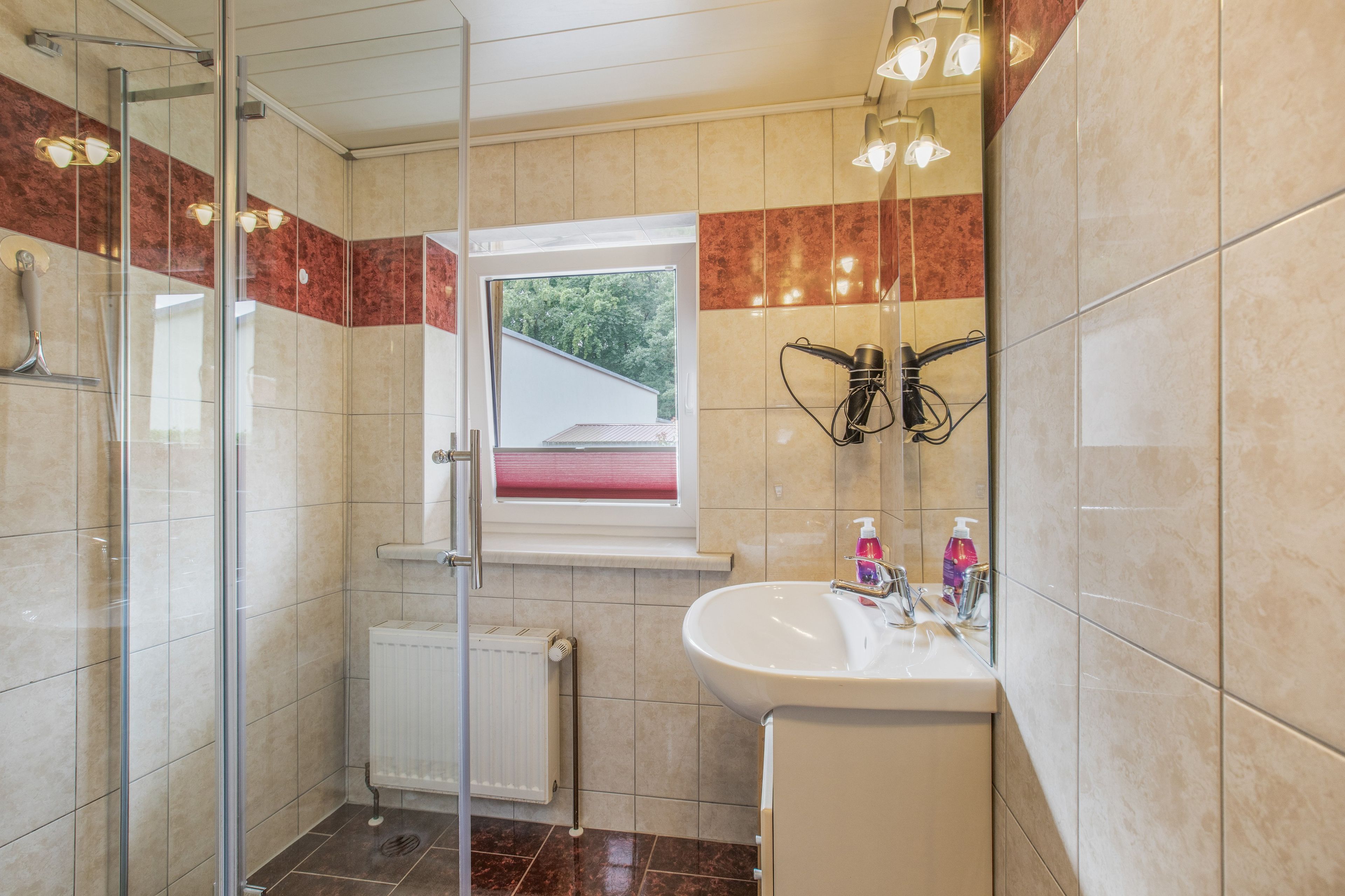 Ein Badezimmer mit Duschkabine aus Glas, einem weißen Waschbecken mit Spiegel und einem Fenster mit Blick auf Grünfläche.