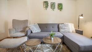 A modern living room with a grey sectional sofa, two round wooden side tables, a grey armchair, and a fluffy white throw pillow. - Apartment “Max” with Private Terrace, Garden, and Wi-Fi (Heringsdorf)