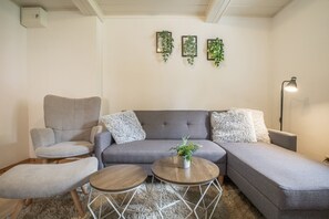 A modern living room with a grey sectional sofa, two round wooden side tables, a grey armchair, and a fluffy white throw pillow. - Apartment “Max” with Private Terrace, Garden, and Wi-Fi (Heringsdorf)