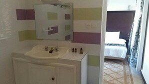 A bathroom with a sink, mirror, and toiletries. There is a bed with a white bedspread and a dark headboard in the adjacent room.