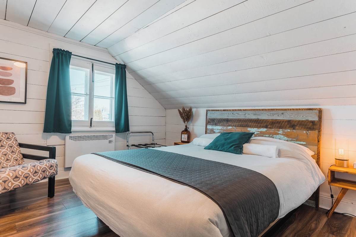 Een slaapkamer met een groot bed, een stoel, een raam met gordijnen, een nachtkastje met een lamp en een muur met een gestructureerd kunstwerk.
