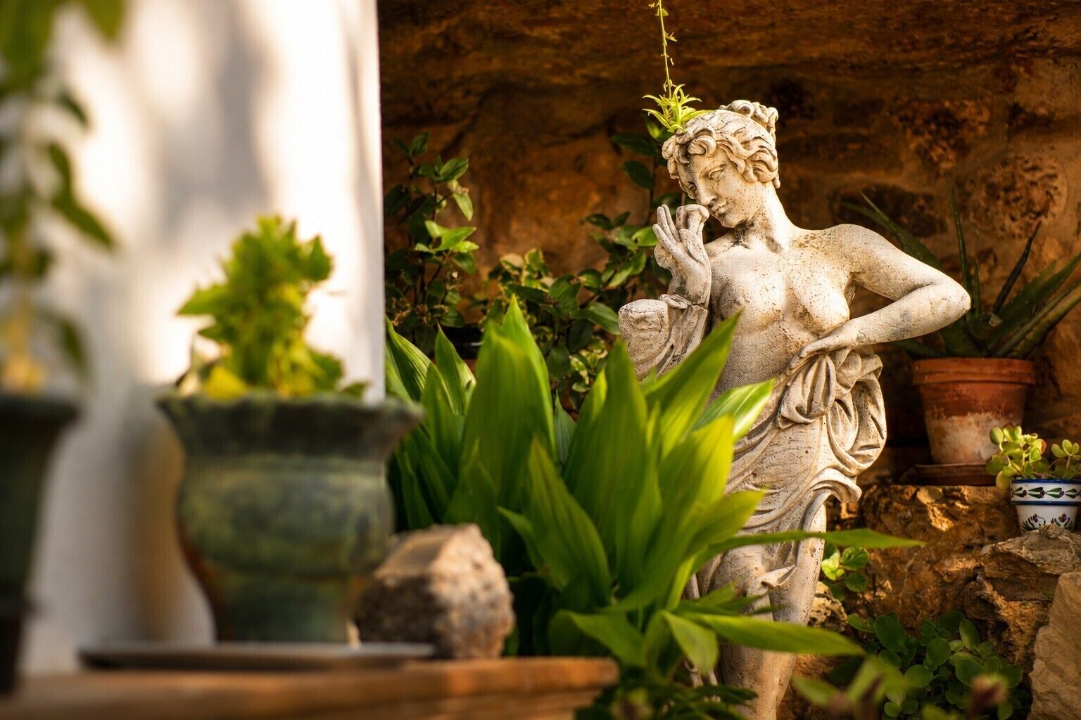 A statue in a garden with potted plants and a rustic stone wall.