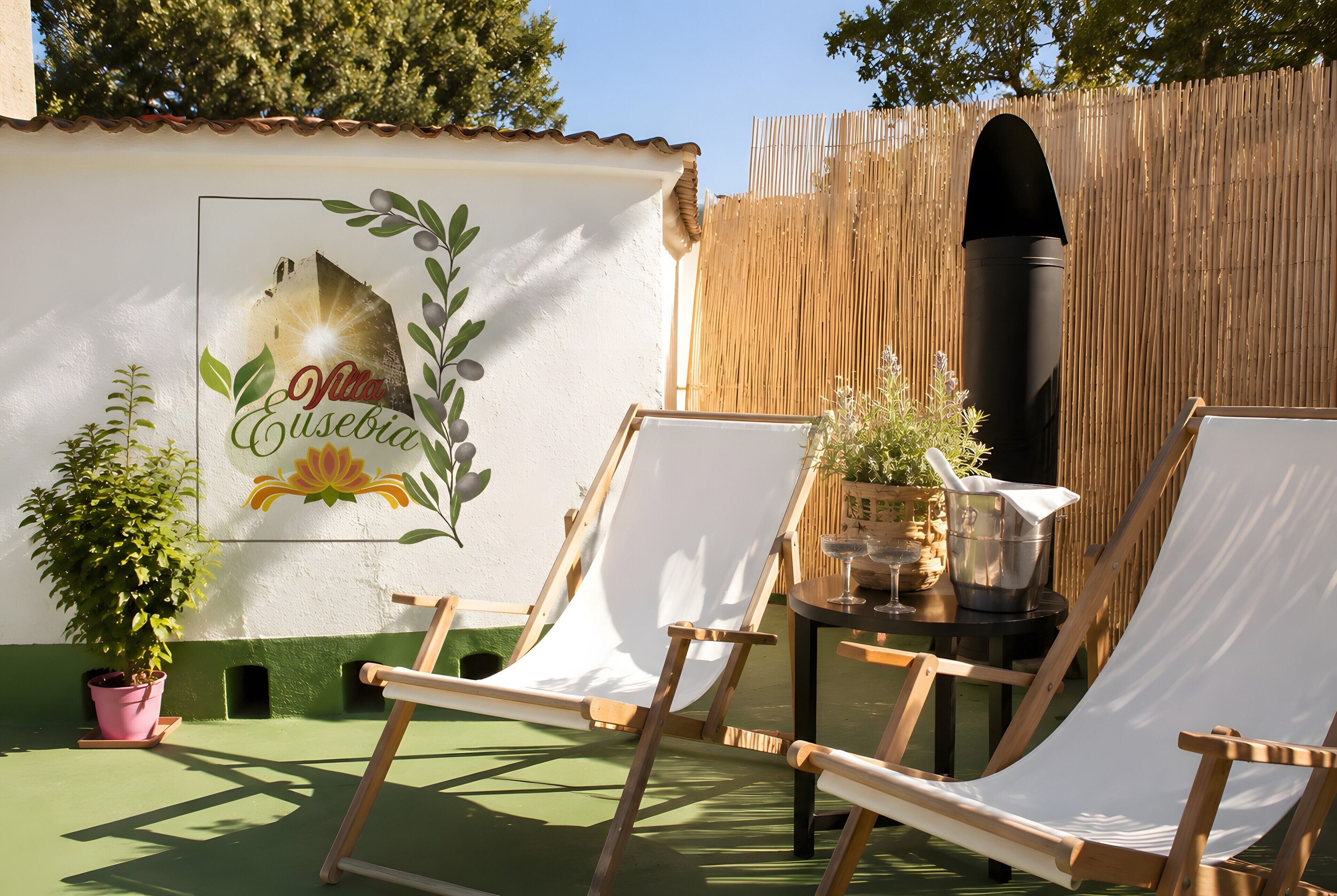 Un patio con dos sillas blancas de lona, una mesa con un balde de metal y copas de vino, y una pared con un mural.
